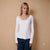 A woman modeling a white long-sleeve knit top with a delicate lace-trimmed scoop neckline, paired with dark blue jeans, set against a solid warm beige studio background.