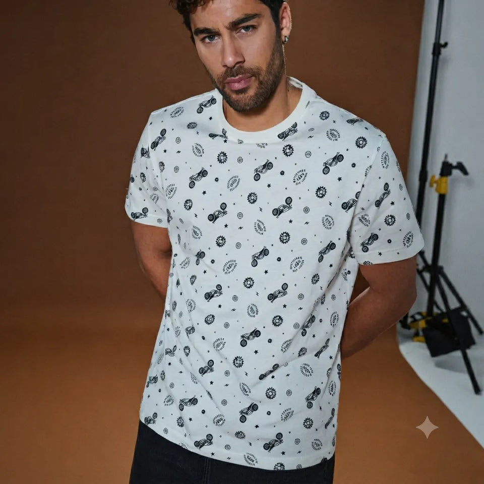 A man wearing a white short-sleeve crewneck t-shirt with a black and gray all-over abstract pattern, posed in a studio setting with a warm brown backdrop.