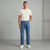 A man wearing a cream t-shirt, blue jeans, and white sneakers stands against a textured gray concrete wall.