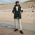 A man stands on a sandy beach wearing a black puffer vest over a white and dark green long-sleeve shirt, off-white pants, and black sneakers.