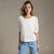 A woman modeling a white textured short-sleeve blouse with sheer puff sleeves and light blue jeans, standing against a neutral beige studio background.