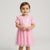 A toddler girl wearing a pink short-sleeve dress with ruffled shoulders and a tulle skirt, featuring a heart-shaped applique on the chest, photographed against a light gray studio background.