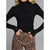 A woman wearing a fitted black long-sleeve turtleneck top with small gold button details on the cuffs, paired with leopard-print pants, against a plain gray studio background.