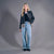 A blonde model stands against a gray studio background wearing a black overshirt over a dark top, a plaid shirt tucked into light-wash wide-leg jeans, and black shoes.