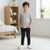 A young boy standing in a bright, modern bedroom wearing a light gray crewneck t-shirt, dark slim-fit jeans, and white sneakers.