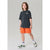 A child model wearing an oversized dark charcoal gray t-shirt with a small white logo on the left chest, paired with bright orange shorts, white socks, and green and white sneakers, standing against a light gray studio background.