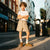 A young boy stands on a city street wearing a white short-sleeve t-shirt, beige drawstring shorts, white socks, and brown sneakers with green stripes. The background shows a blurred urban street scene with buildings and warm sunlight.