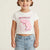 A young girl with her hair in pigtails smiling, wearing a white short-sleeve crop top t-shirt with a pink 'PARADISE' graphic and light blue jeans, set against a plain off-white background.