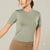 A woman wearing a fitted short-sleeve crewneck athletic top in sage green, paired with tan leggings, against a plain light gray studio background.