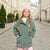 A young girl with long braids stands on a cobblestone street wearing an oversized sage green parka jacket, a pink knit beanie, and pink pants.
