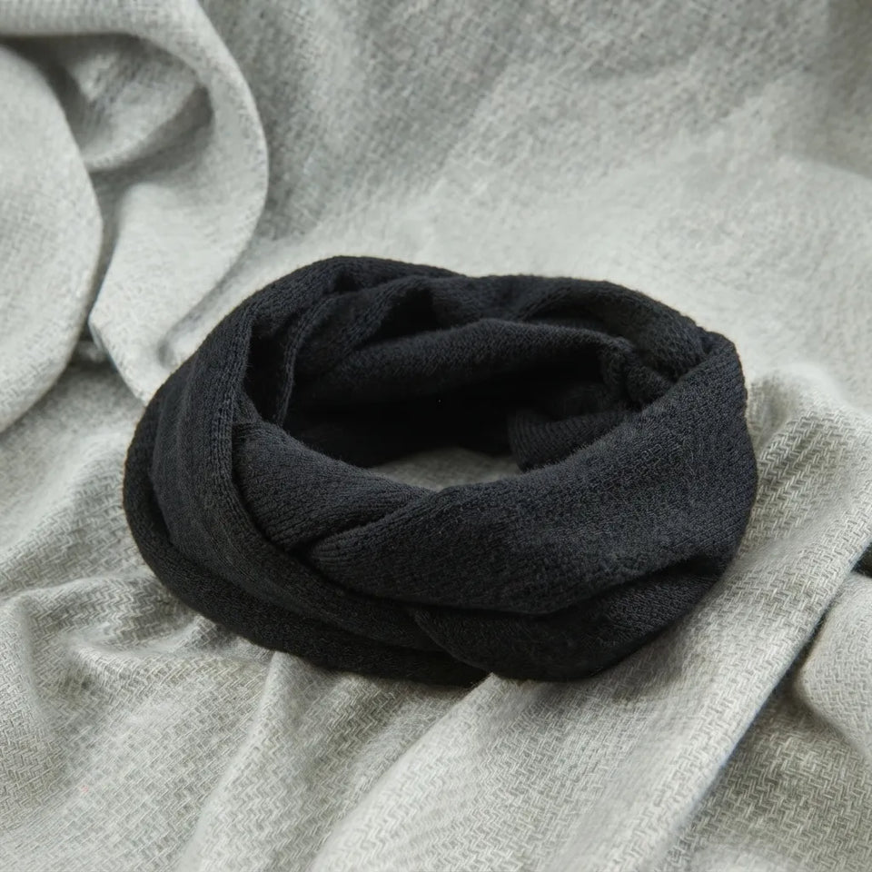 A black infinity scarf made of soft, textured fabric, styled on a light gray draped background.