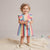 A smiling toddler girl stands on a cream knit blanket in a studio setting, wearing a pastel rainbow striped dress with ruffled shoulders and holding a small plush toy. No visible brand markings.