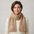 A woman wearing a beige cable-knit cowl scarf with a gold embroidered monogram on the left side, paired with a cream sweater against a light gray studio background.