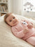A baby lying on a cream-colored textured blanket in a softly lit nursery, wearing a light pink long-sleeve footed sleeper with a black and white panda face patch on the chest.