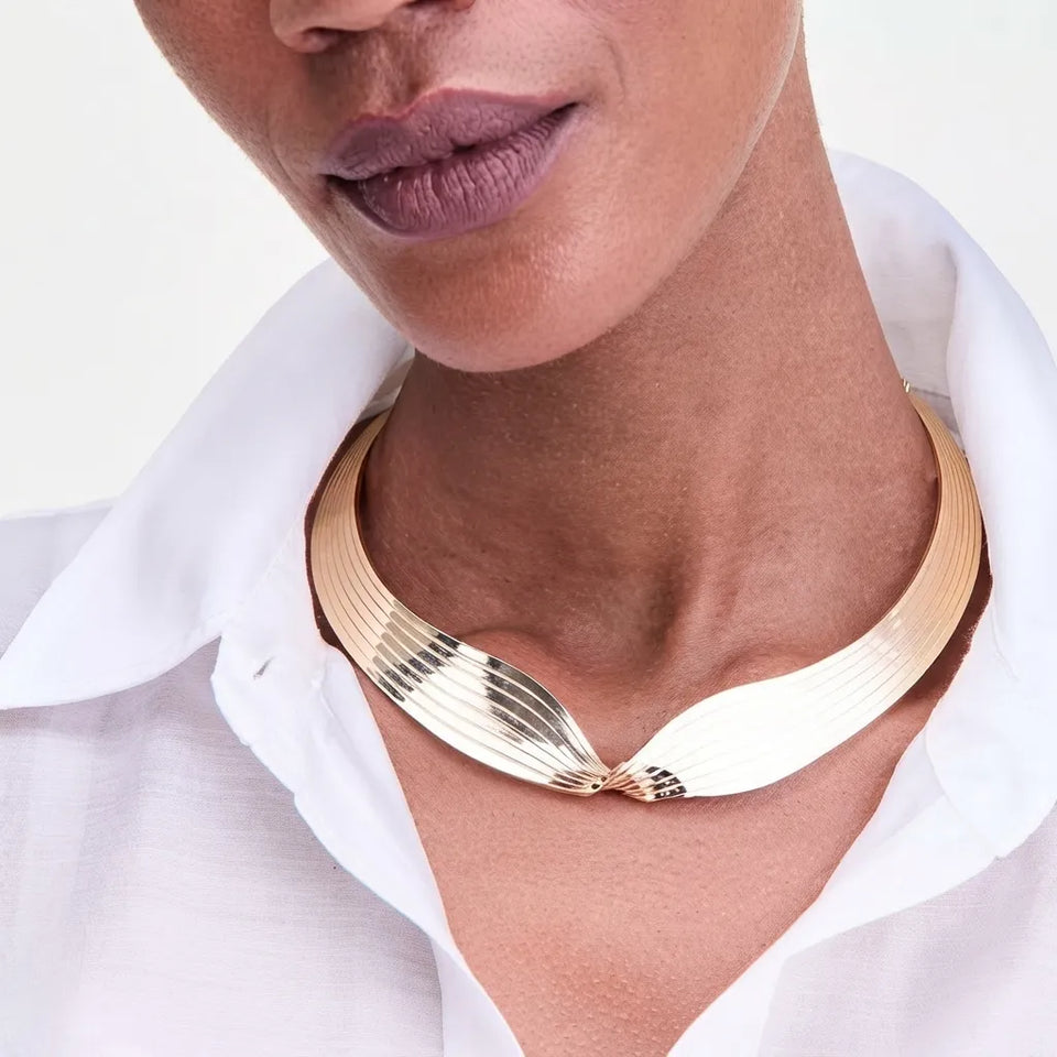 A close-up of a model wearing a wide, chunky gold-toned collar necklace with a textured, ribbed design and a twisted knot detail at the front, styled with a white collared shirt.