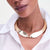 A close-up of a model wearing a wide, chunky gold-toned collar necklace with a textured, ribbed design and a twisted knot detail at the front, styled with a white collared shirt.