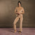 A woman stands barefoot against a textured brown backdrop wearing a matching beige lounge set consisting of a long-sleeve cropped top and wide-leg pants.