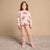 A young girl stands against a plain beige studio backdrop wearing a matching two-piece pajama set. The set is light pink with a brown bunny print on the long-sleeve top and matching pink pants featuring bunny ears and bunny faces on the knees.