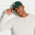 A man wearing a dark green baseball cap with a white letter 'A' and a cream-colored long-sleeve henley shirt, posing against a white brick wall.