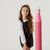 A young girl with long wavy brown hair stands against a light gray studio background, holding a large pink pencil prop. She is wearing a dark brown leopard-print pinafore dress over a white long-sleeve shirt with small blue floral prints.