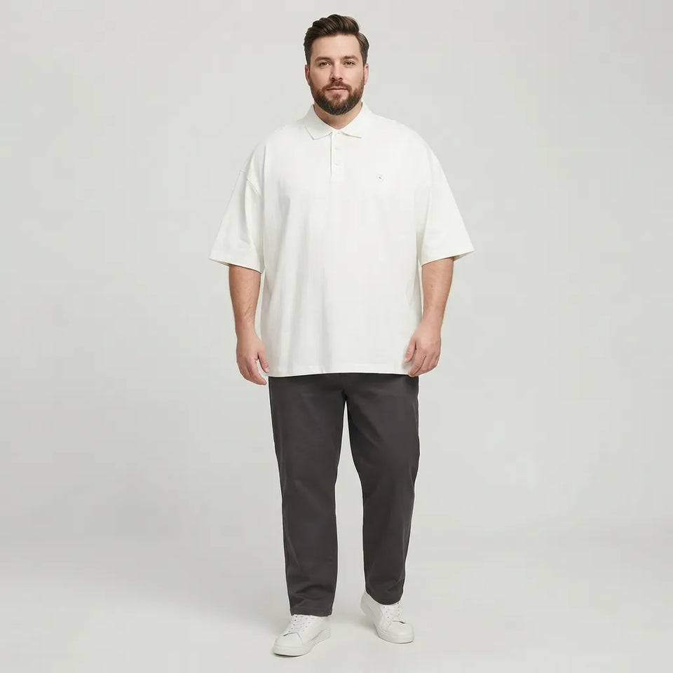 A man modeling an oversized white short-sleeve polo shirt paired with dark gray pants and white sneakers, standing against a light gray studio background.