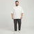 A man modeling an oversized white short-sleeve polo shirt paired with dark gray pants and white sneakers, standing against a light gray studio background.