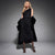 A woman modeling a black one-shoulder midi dress with ruched detailing, styled with a black velvet coat draped over her arms, set against a dark gray studio background.