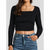A woman wearing a black long-sleeve crop top with a square neckline and blue jeans, shown from the neck down against a light gray background.