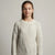 A young girl with light brown hair tied back, wearing a cream-colored cable-knit crewneck sweater, photographed against a neutral light gray studio background.
