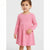 A young girl wearing a pink long-sleeve dress with a white polka dot pattern, standing against a light gray studio background.