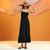 A woman modeling a black spaghetti-strap maxi dress with a tiered ruffle hem, styled with a wide-brimmed straw hat and black strappy sandals, set against a warm orange abstract background.