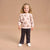 A young girl with curly blonde hair stands against a solid beige studio background, wearing a light pink sweatshirt with a cartoon monkey print, dark gray pinstriped pants, and white sneakers.