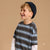A young boy smiling and looking to the side, wearing a dark navy knit beanie and a dark gray striped t-shirt with blue and white stripes over a white long-sleeve shirt, set against a solid beige background.