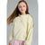 A young girl with long brown hair wearing a pale yellow-green hooded sweatshirt with a small embroidered logo on the chest, paired with pink and white striped pants, set against a plain gray studio background.