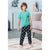 A young boy stands barefoot on a white rug in a living room setting, wearing a short-sleeve mint green t-shirt with a small circular graphic on the left chest and black pajama pants covered in a white soccer ball pattern.