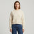 A woman wearing a cream-colored crewneck knit sweater and dark blue jeans, standing against a light gray studio background.
