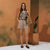 A woman stands in a studio setting wearing a sleeveless top with a gray, black, and beige abstract pattern, paired with light beige shorts. She holds a woven straw bag and wears black flat shoes, set against a warm terracotta-colored background with a potted plant on a pedestal.