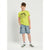 A young boy modeling a bright lime green t-shirt with a black dinosaur graphic and text, paired with light blue denim shorts, white socks, and green and white sneakers, standing against a light gray studio background.