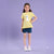 A young girl stands against a solid lavender studio background wearing a pale yellow ruffled short-sleeve t-shirt with a white llama graphic, blue denim shorts, pink socks, and white sneakers.