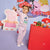 A young child wearing a light pink Peppa Pig themed lounge set stands in a colorful studio setting, pointing toward a large character cutout. The set includes a matching long-sleeve top and shorts with a ruffled hem, and the background features Peppa Pig artwork and a red display table with toy figures.