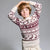 A young boy with light brown hair wearing a cream and burgundy Fair Isle knit sweater with reindeer patterns, posing with his arms raised against a solid gray studio background.