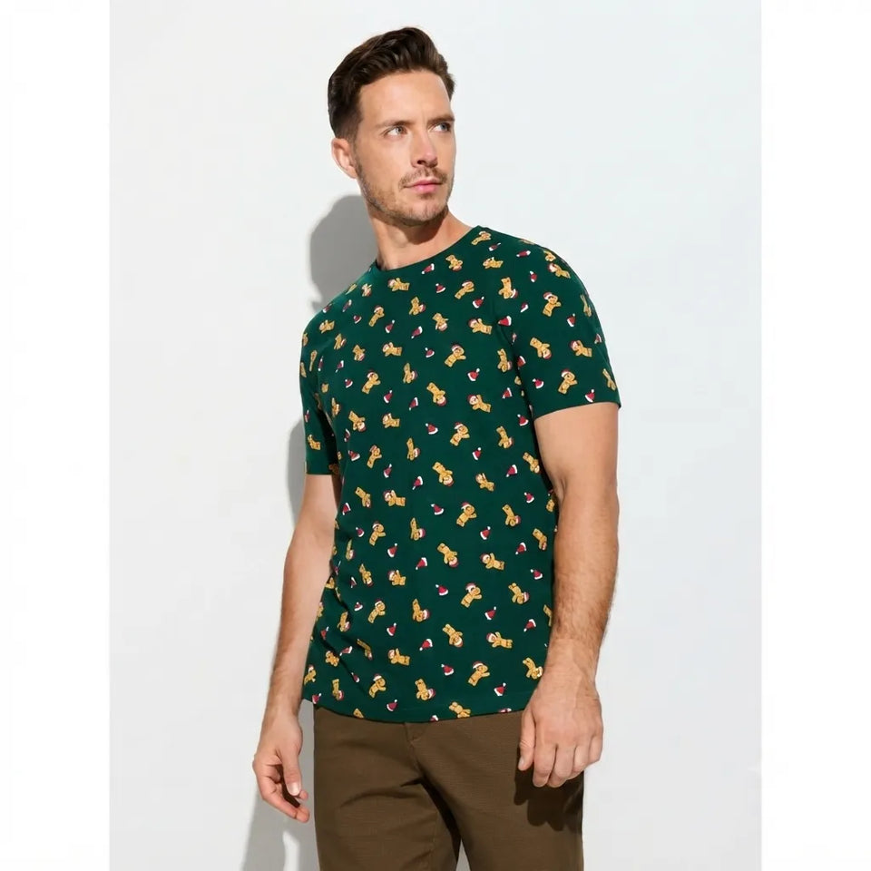 A man wearing a dark green short-sleeve t-shirt with a repeating pattern of small yellow cartoon figures, paired with brown pants, standing against a light gray studio background.