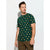 A man wearing a dark green short-sleeve t-shirt with a repeating pattern of small yellow cartoon figures, paired with brown pants, standing against a light gray studio background.