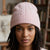 A close-up portrait of a woman wearing a light pink cable-knit beanie hat, with a softly blurred indoor background featuring bookshelves.