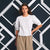 A woman modeling a white short-sleeve blouse with textured puff sleeves and a round neckline, paired with beige wide-leg pants, set against a dark blue and white geometric paneled wall.