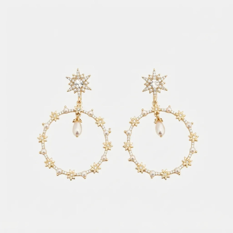 A pair of gold-toned statement dangle earrings featuring starburst studs, small pearl-like beads, and circular hoops accented with tiny clear stones, shown on a light gray background.