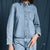 A person wearing a light blue denim button-up shirt with a chest pocket and matching denim bottoms, photographed against a dark blue fabric backdrop.