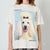 A person wearing an oversized off-white graphic T-shirt featuring a large printed portrait of a fluffy white poodle with a black bow on a light blue background, with black text around the print.