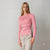 A woman modeling a pink long-sleeve ribbed knit top with an asymmetrical draped design and gathered side detail, paired with light gray trousers, against a plain light gray studio background.