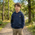 A young boy stands on a gravel path in a wooded outdoor setting, wearing a navy blue quarter-zip hoodie with black elbow patches and a small circular chest logo, paired with khaki cargo pants.
